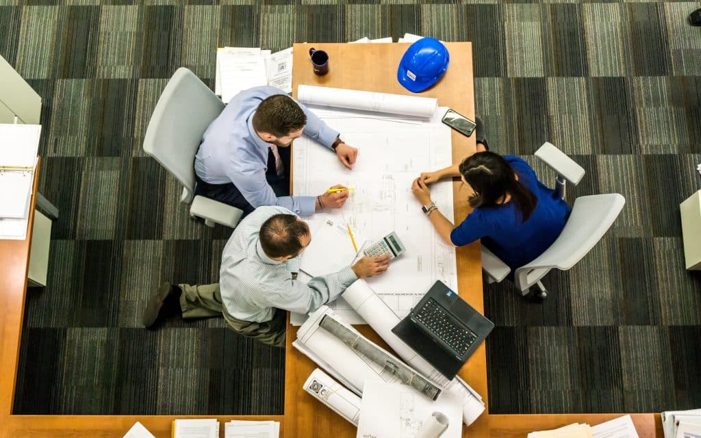 Two males and one female sat at an office desk with paper and a laptop working together